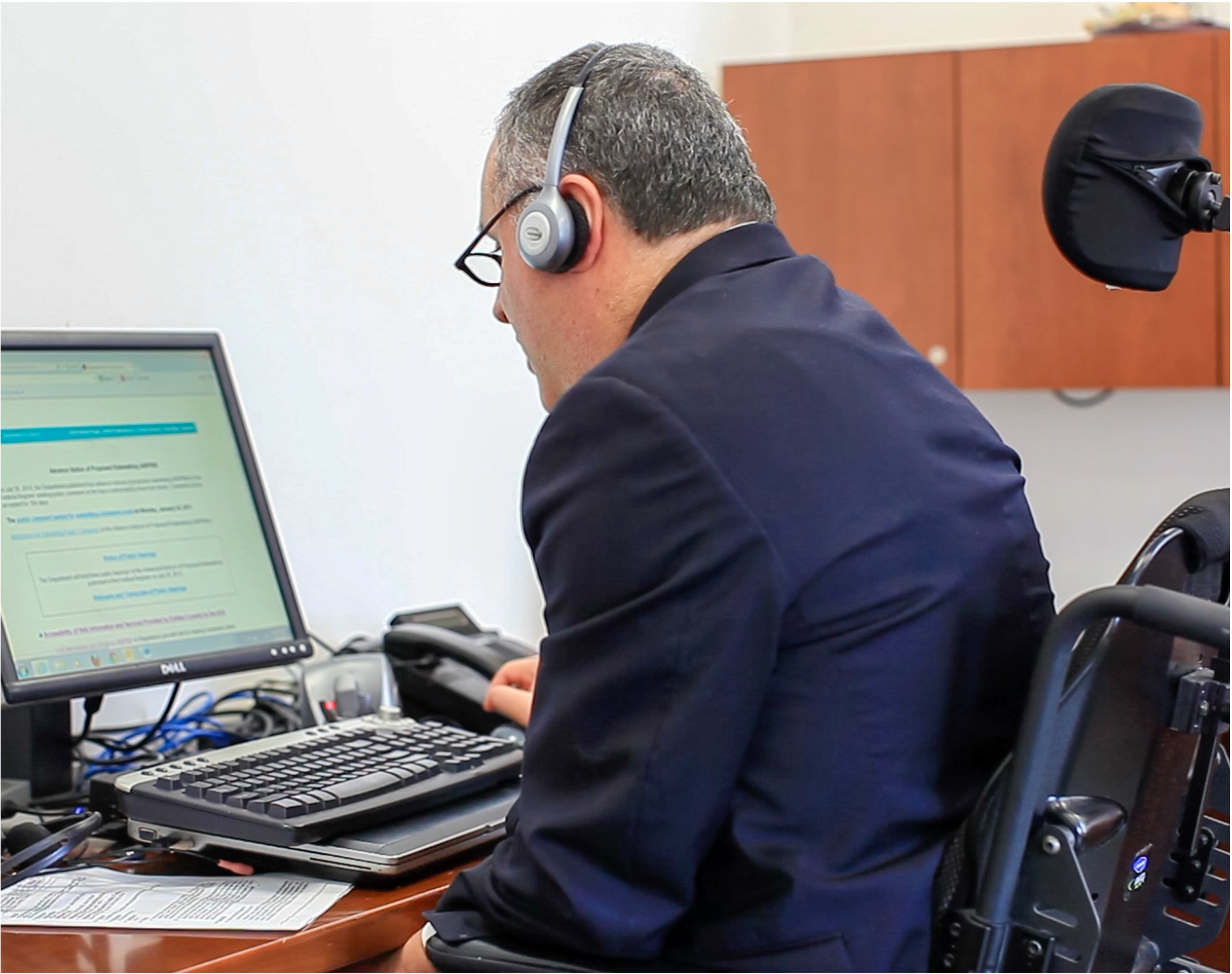 A wheelchair user wearing headphones and listening to website content with assistive technology. They are reading along with the content on their computer monitor.